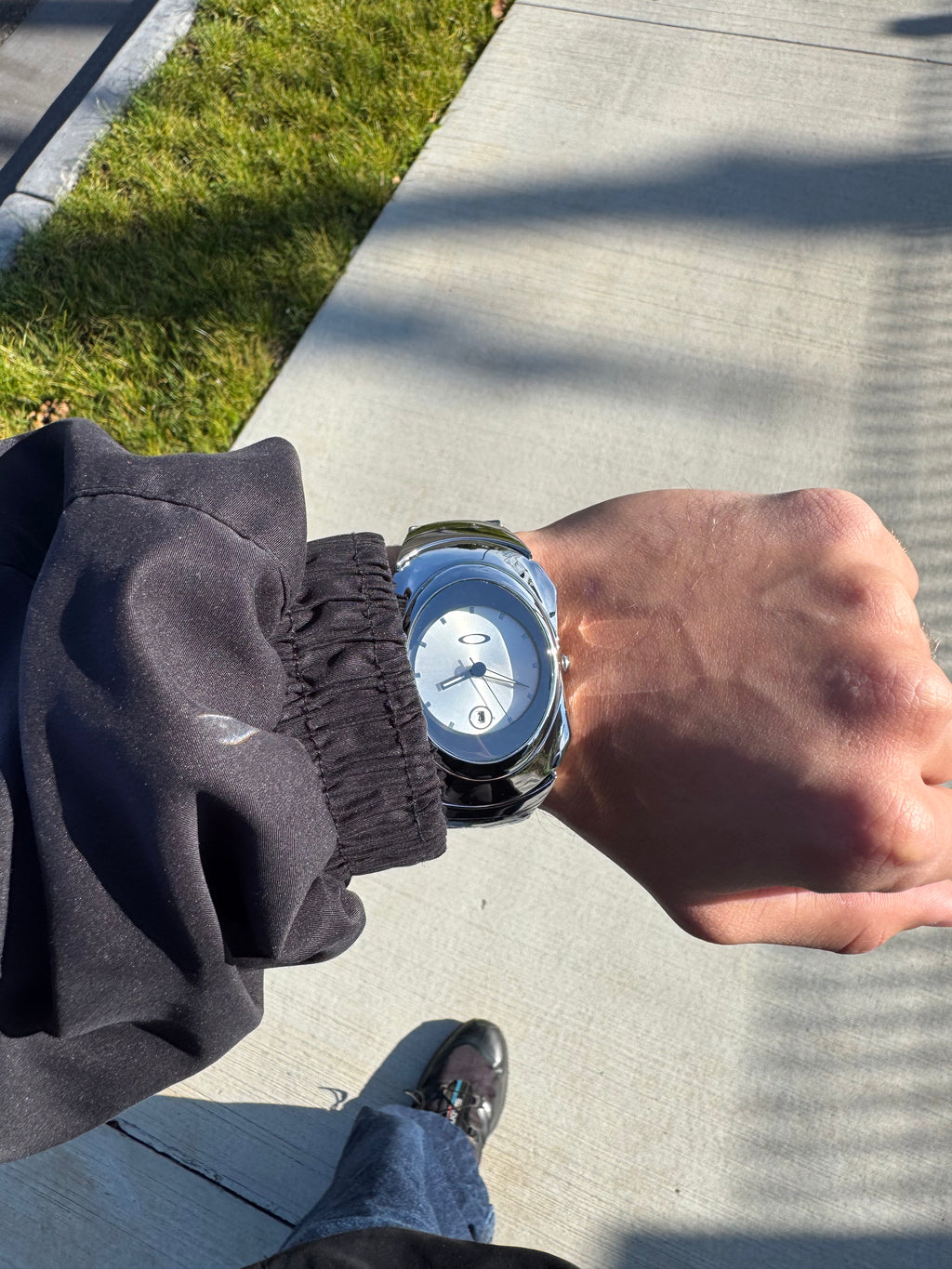 Top-down lifestyle view of chrome Y2K watch worn on wrist over light concrete pavement, Aerostore Glossy Ice White showing Frutiger Aero polished stainless steel sculptural case in natural daylight