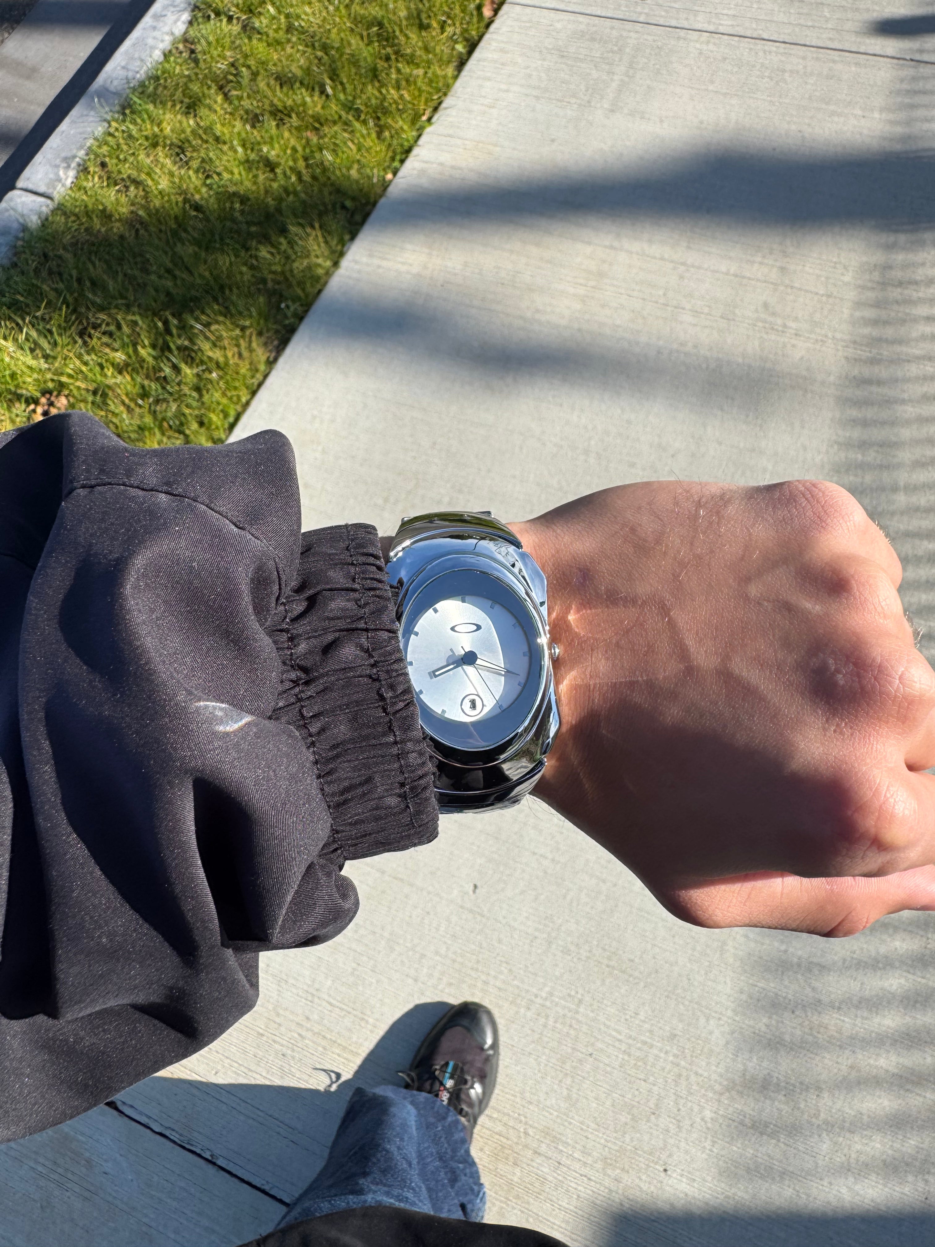 Top-down lifestyle view of chrome Y2K watch worn on wrist over light concrete pavement, Aerostore Glossy Ice White showing Frutiger Aero polished stainless steel sculptural case in natural daylight