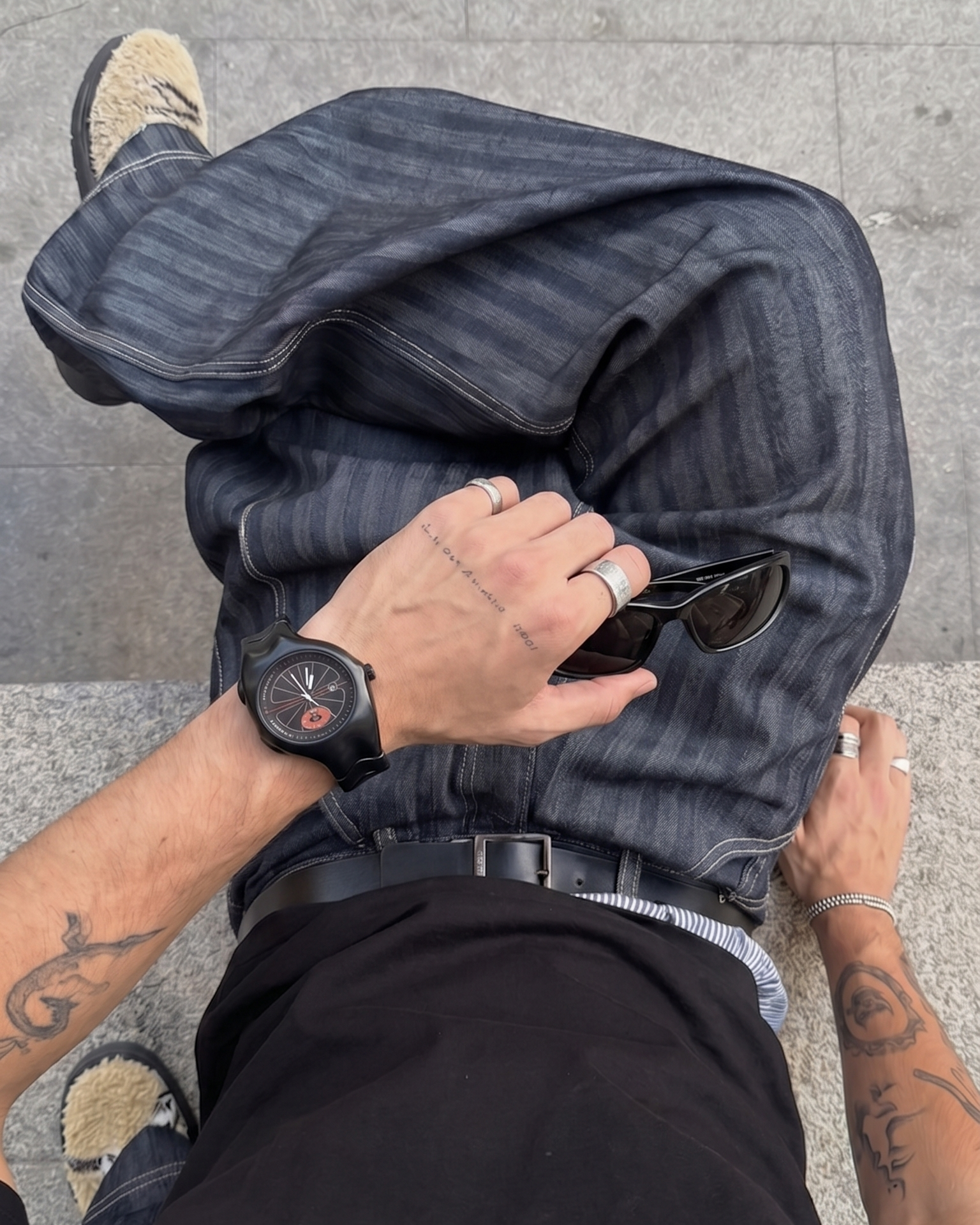 Person wearing a dark suit with a sports y2k watch from the 2000's , also with sunglasses on a gray pavement background