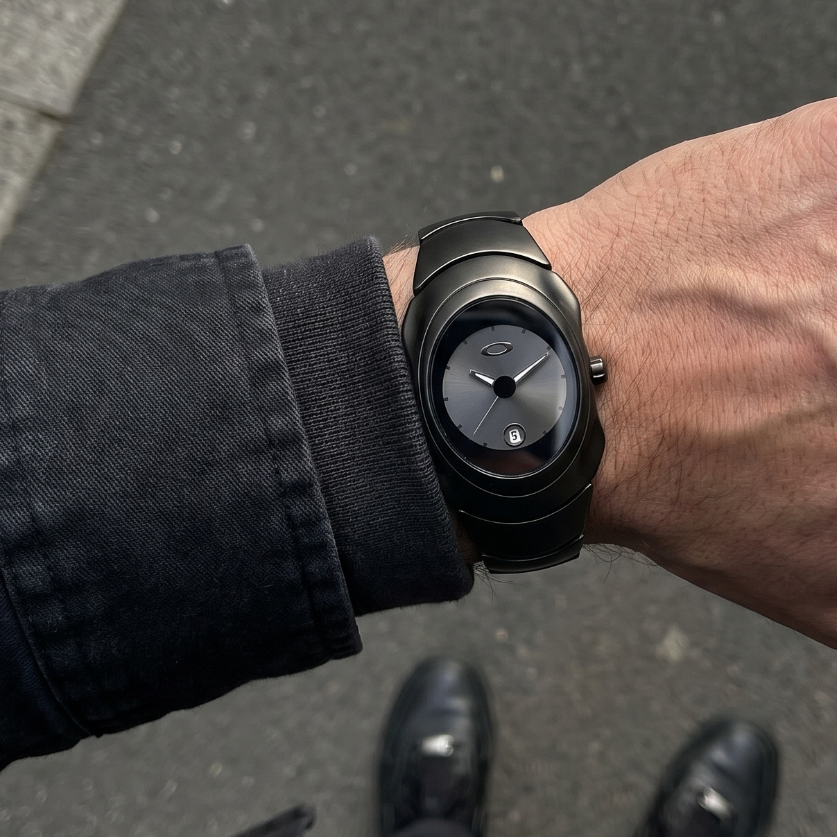 Y2K-style black watch worn on wrist with dark sleeve, top-down lifestyle view on city pavement showing Aerostore Midnight Black early-2000s industrial design