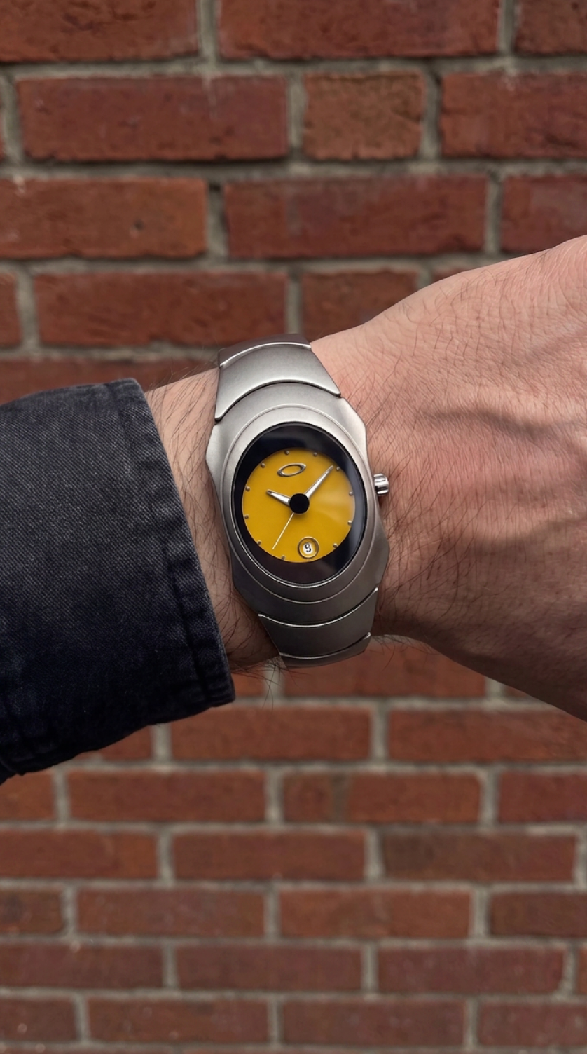 Matte metal Y2K watch with glossy yellow dial worn against red brick wall, Aerostore Matte Yellow showing Frutiger Aero industrial design language and early-2000s sculptural case geometry in urban setting