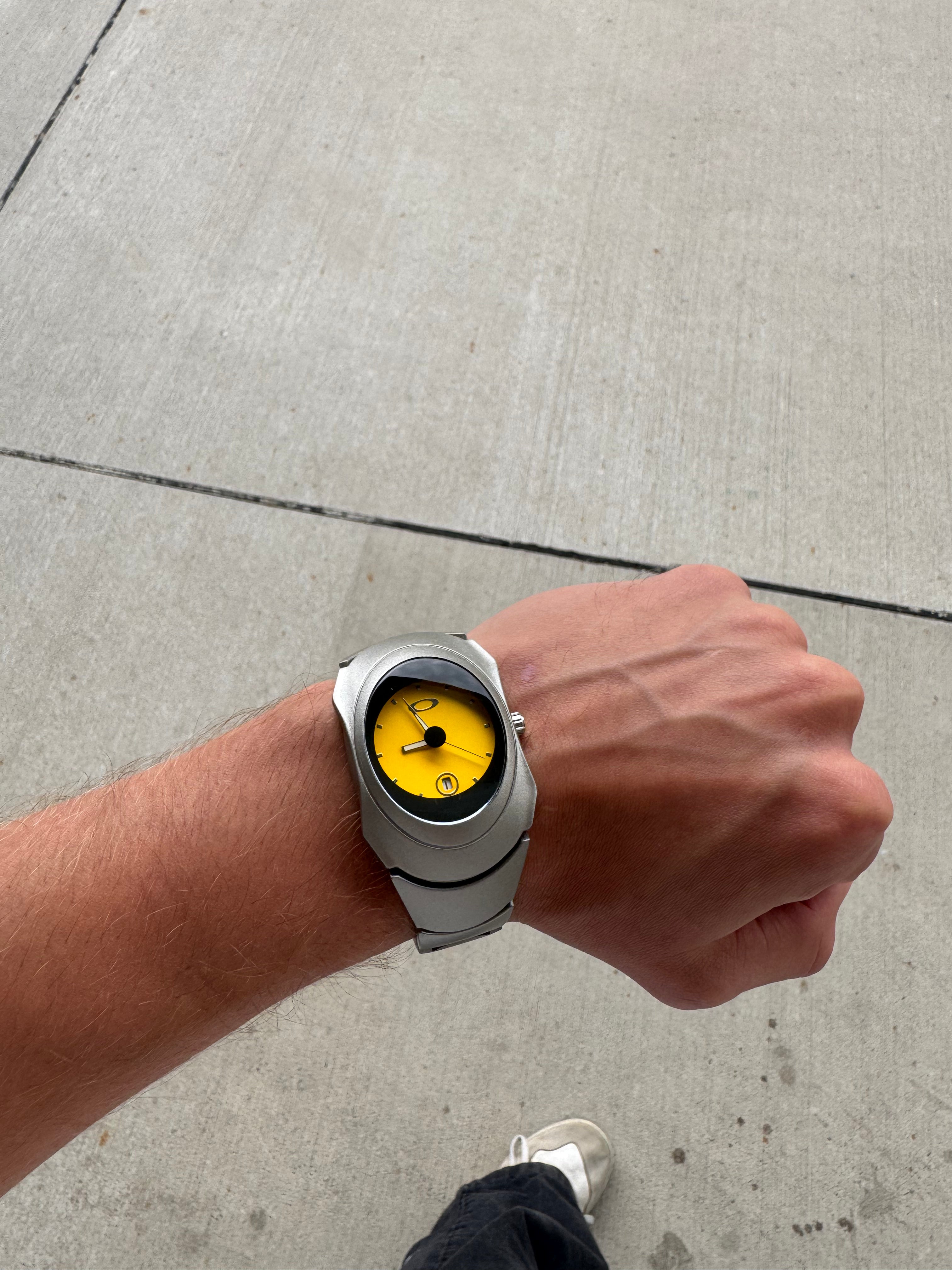 Y2K wristwatch with glossy yellow dial worn casually, Aerostore Matte Yellow showing matte stainless steel finish, Frutiger Aero colour palette and sculptural early-2000s industrial case